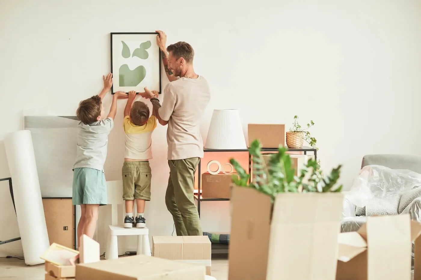 A man with two young boys are trying to hang a framed picture on the wall in their new house. The room is full of moving boxes.