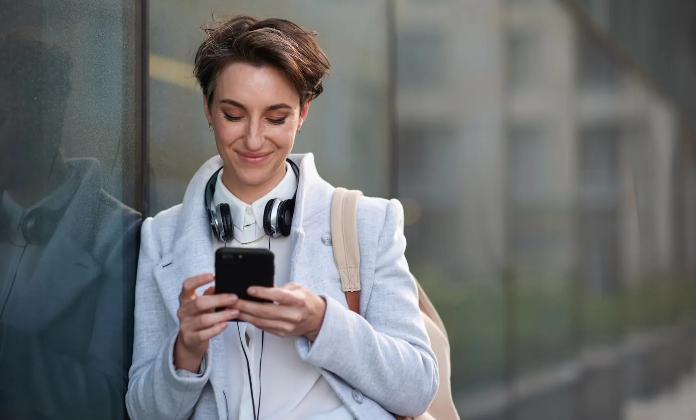 Smiling young woman standing outside an office building, checking her smartphone with headphones resting around her neck