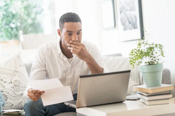 Concerned young man looking at his laptop while holding a printout
