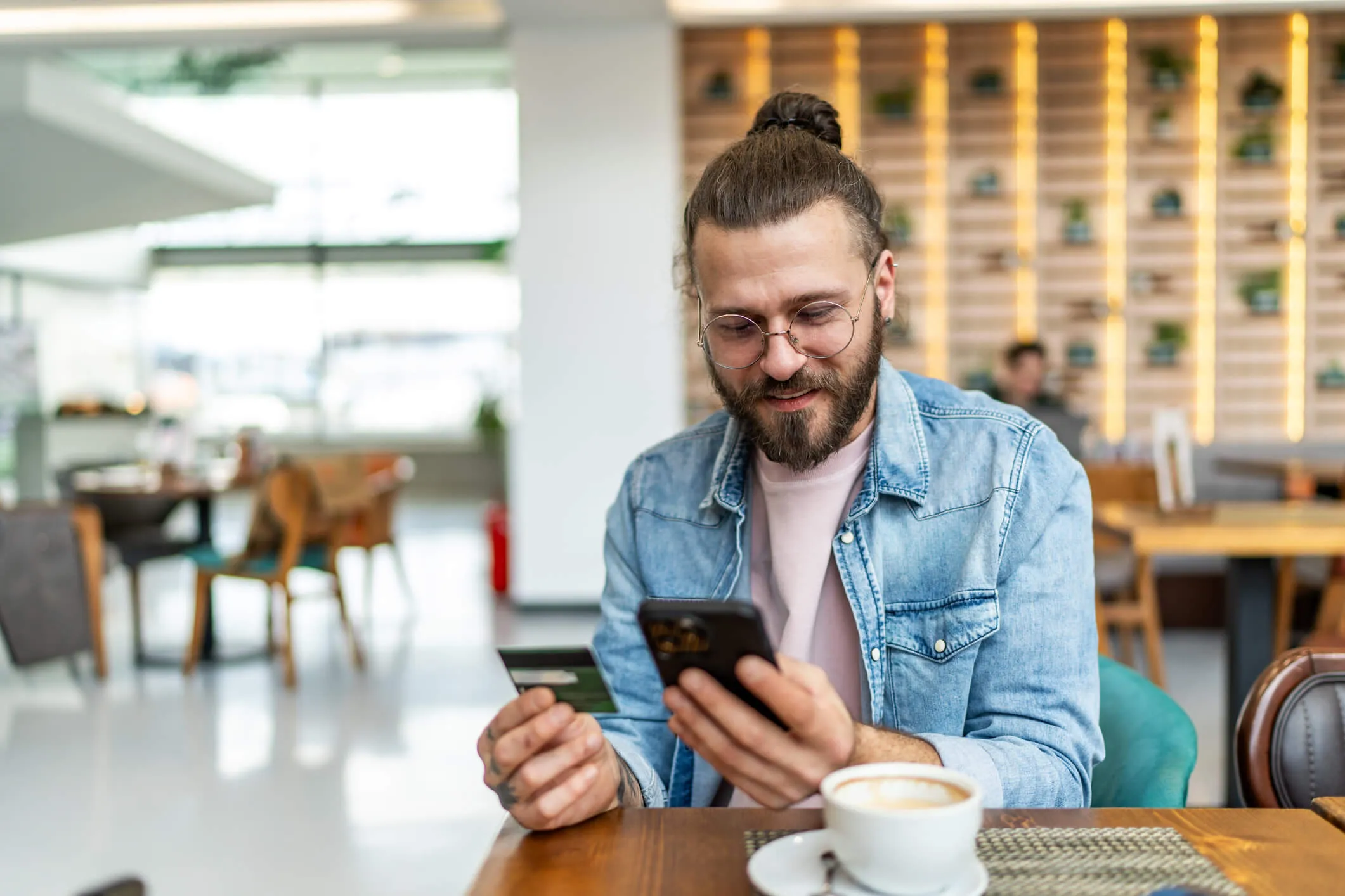 Man sits at a café table holding a credit card and using a smartphone, with a cup of coffee in the foreground.