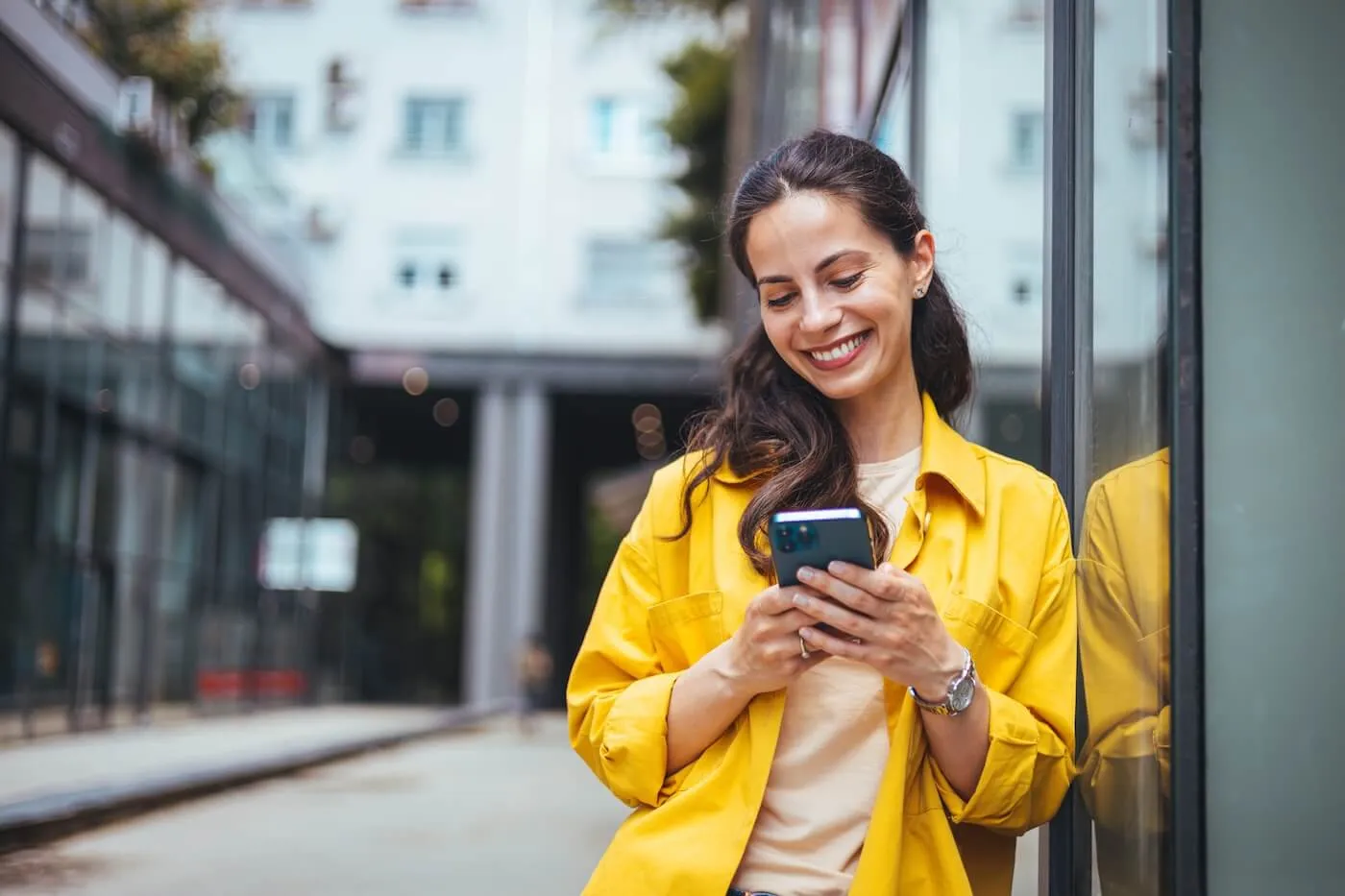 Woman in a yellow jacket smiling while checking her phone in an urban setting