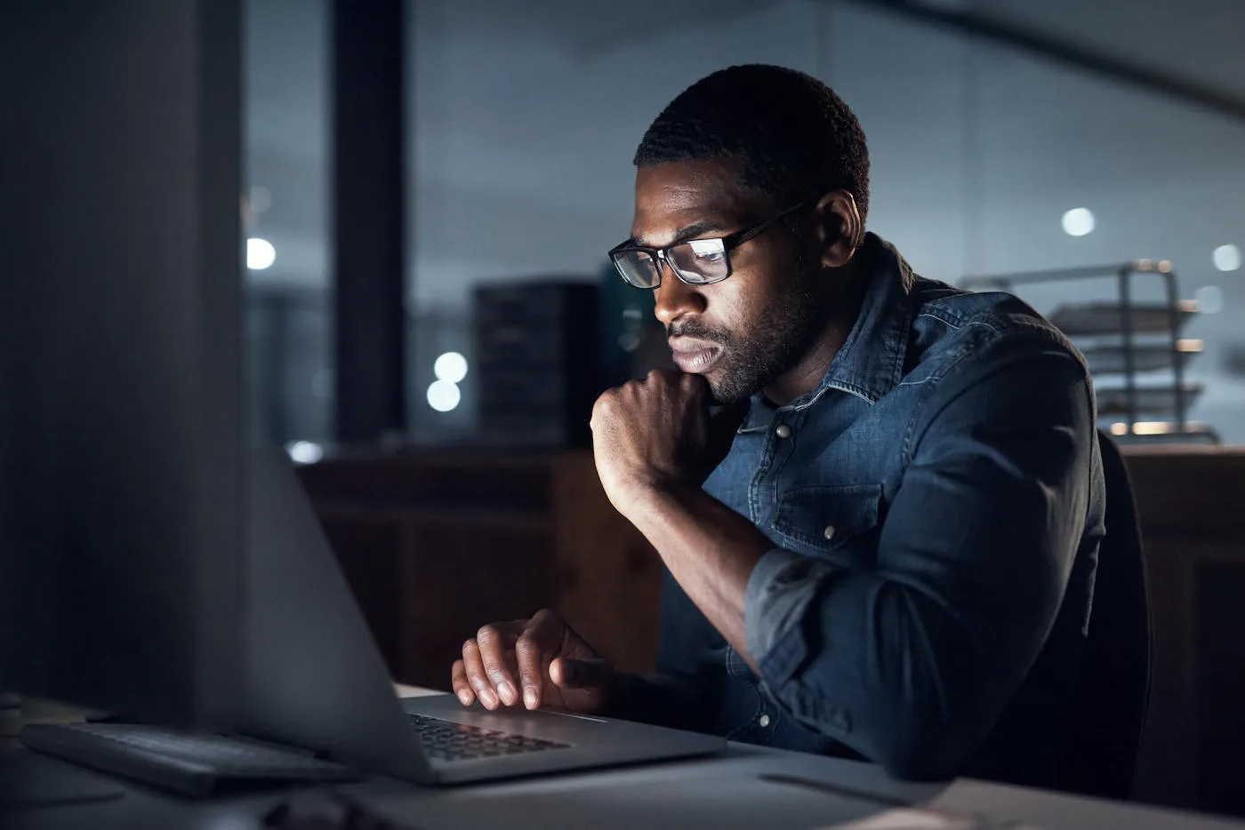 A man wearing a denim shirt and glasses looks at his laptop screen in a dark room.