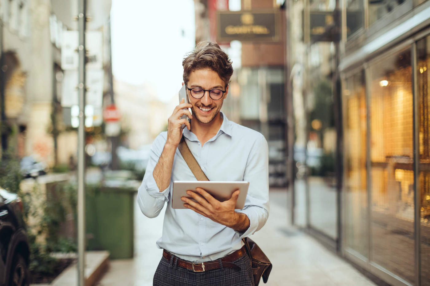 A man wearing glasses talks on the phone while smiling at his tablet outside.