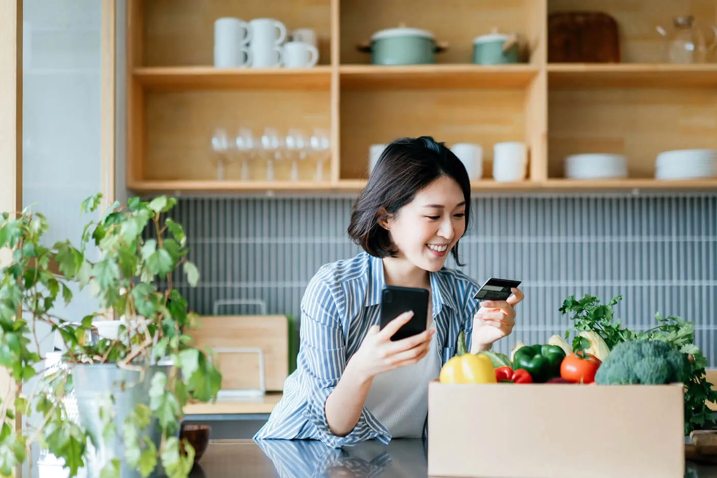 A smiling young woman holding her credit card, with a box of colourful and fresh organic groceries on the kitchen counter at home.