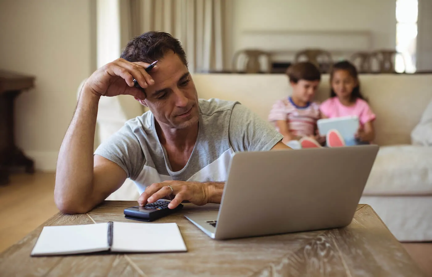 Focused mature man filing taxes online, with two young children sitting on a sofa in the background using a tablet