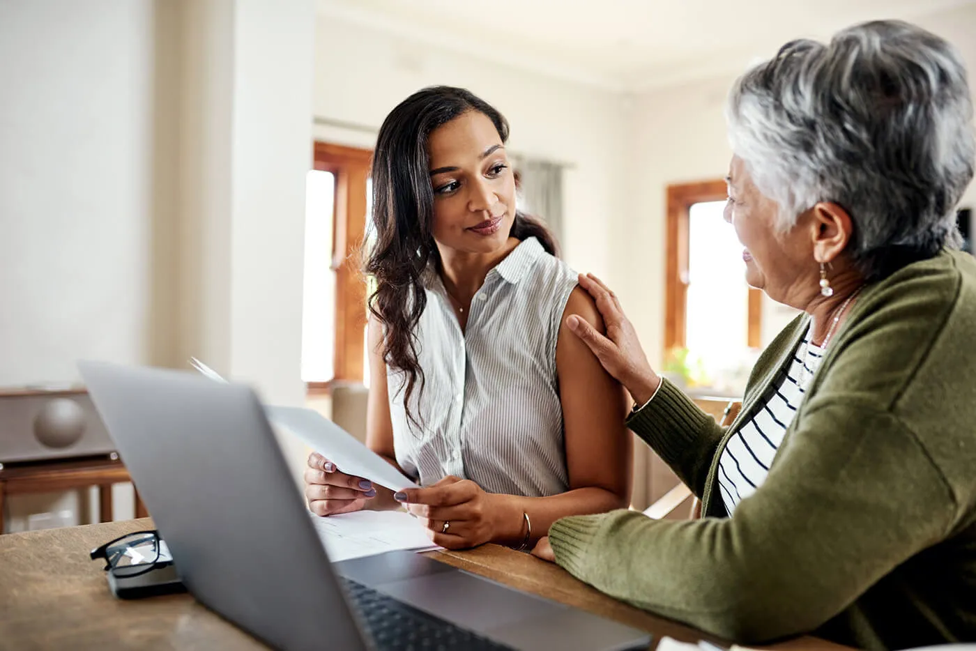 Young woman explaining financial documents to her grandmother at home, with an open laptop beside them
