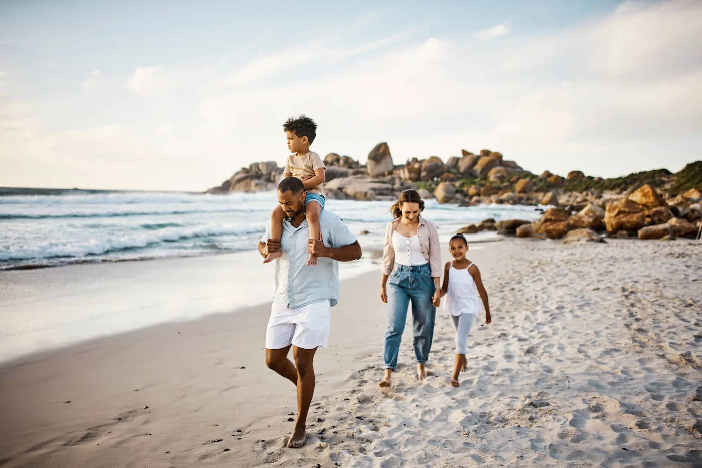 A mother holds the hand of her daughter while father has son carrying him while they walk on the beach.