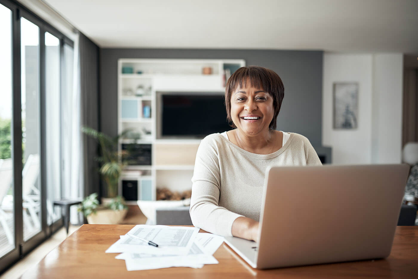 Smiling mature woman using her laptop at home, with the printed documents on the desk