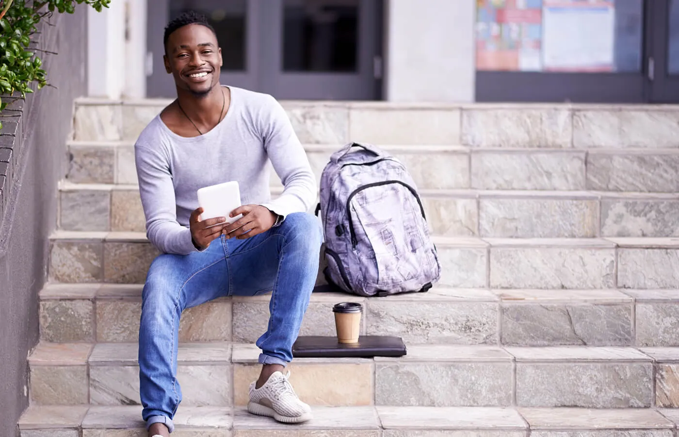 Young man in long sleeve shirt and denim sitting on a step next to a backpack smiling