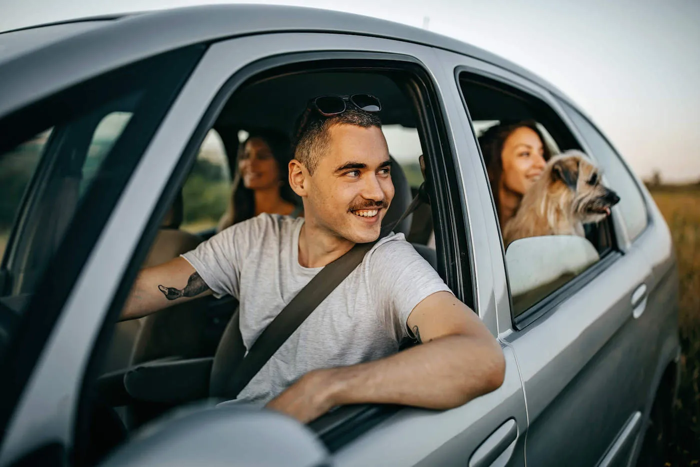 A man smiles while looking out the window of his car with passengers and a dog in the back seat.