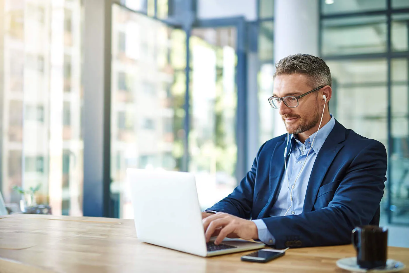 A man wearing a blue suit is using earphones while typing on his laptop at the office.