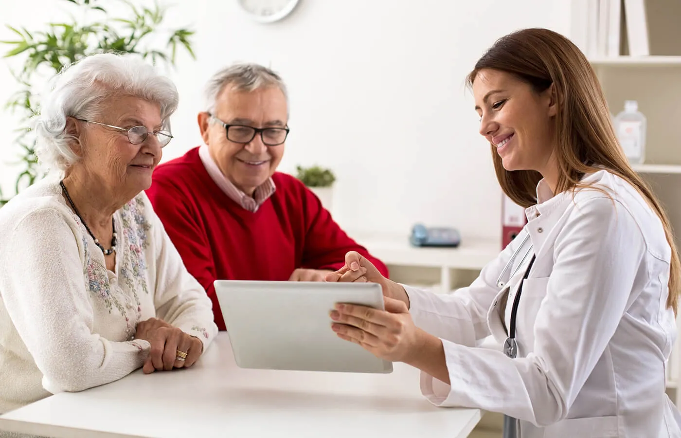 A doctor meets with a smiling elderly couple at a table, reviewing data on an electronic tablet.
