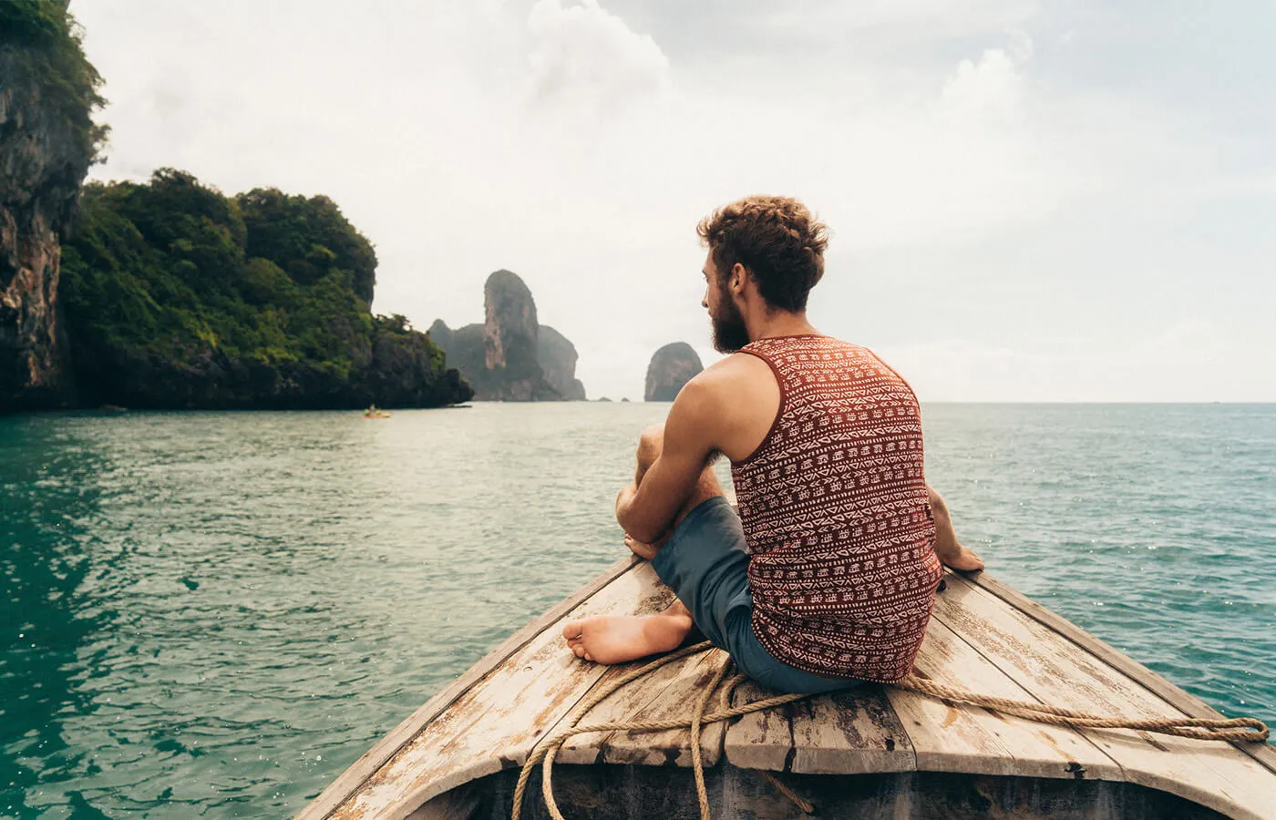 Relaxed man sitting cross-legged on the bow of a wooden boat, facing open water and distant rocky islands, with teal-colored sea surrounding the boat and steep, tree-covered cliffs visible along the shoreline under a light sky.