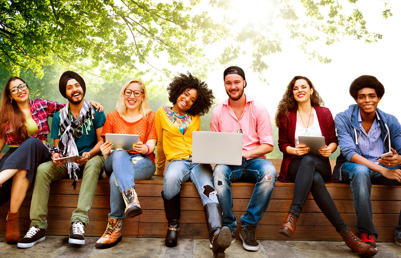 Students sit on an outdoor bench holding laptops and tablets while posing for a group photo.