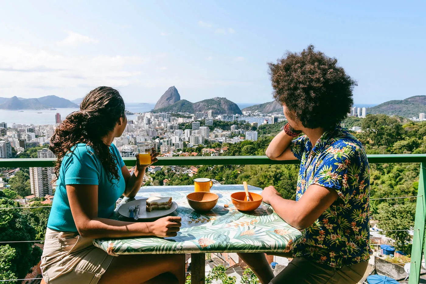 Two people seated at a small outdoor table on a balcony, looking out over a city skyline and coastal landscape, with bowls, plates, drinks, and a patterned tablecloth on the table, and buildings, greenery, and distant hills visible in the background.
