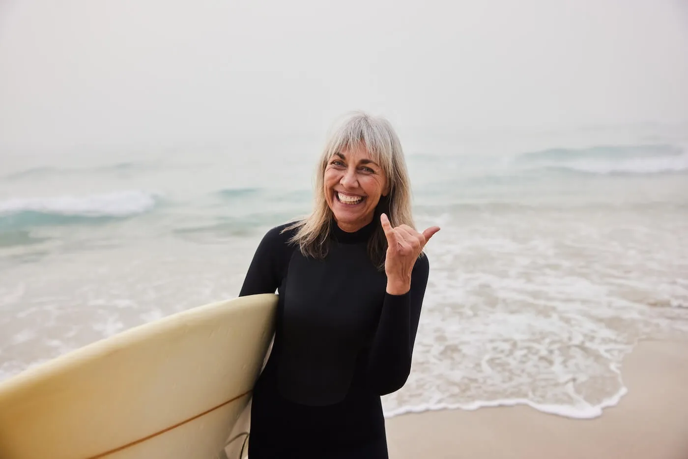 Smiling mature woman in a wetsuit holding a white surfboard and making a shaka sign by the water’s edge