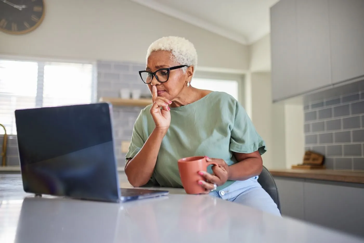 Senior woman using her laptop while having a hot dring in the kitchen
