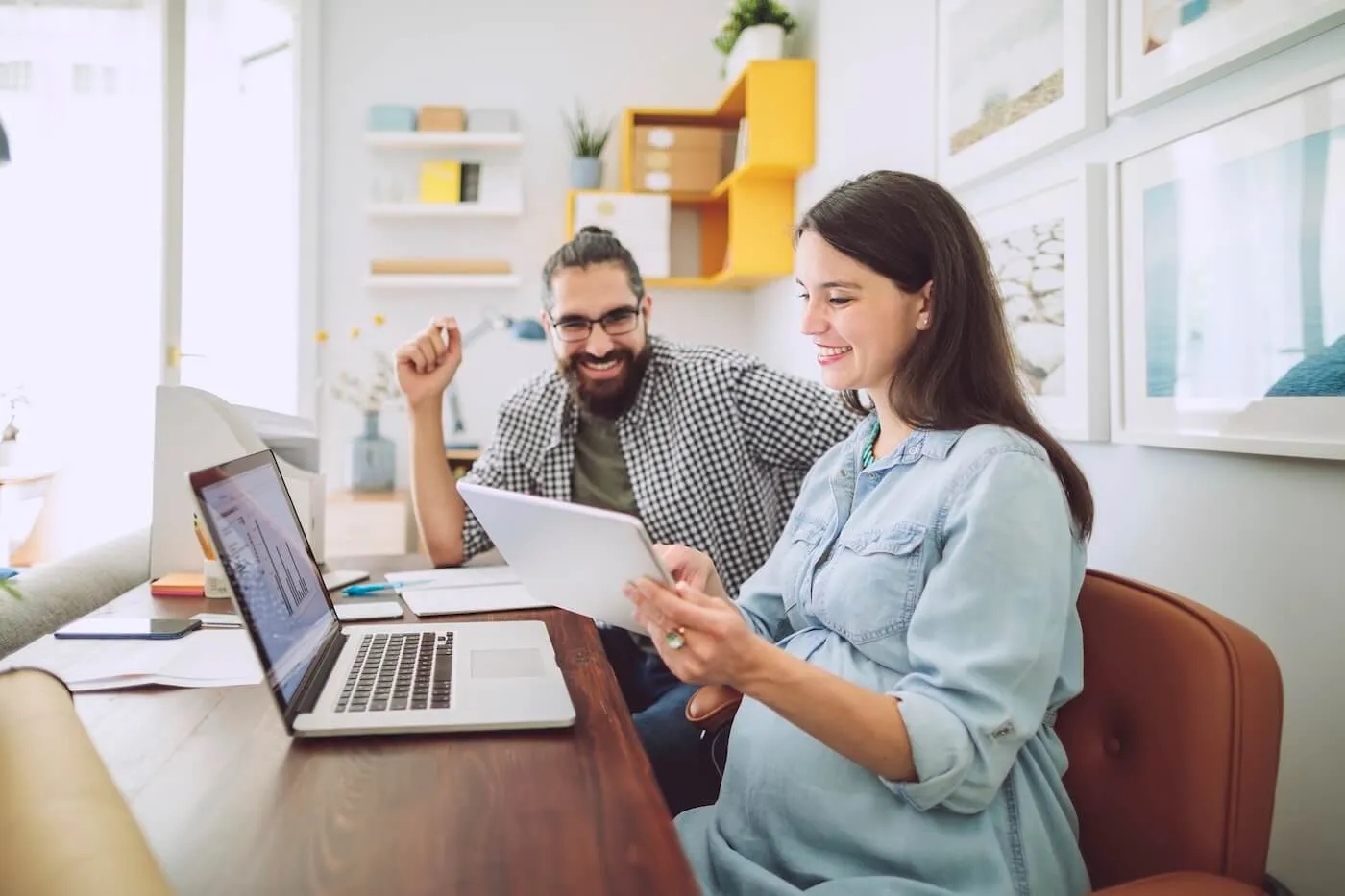 Happy couple discussing finances at home; pregnant woman showing something to her husband on a tablet