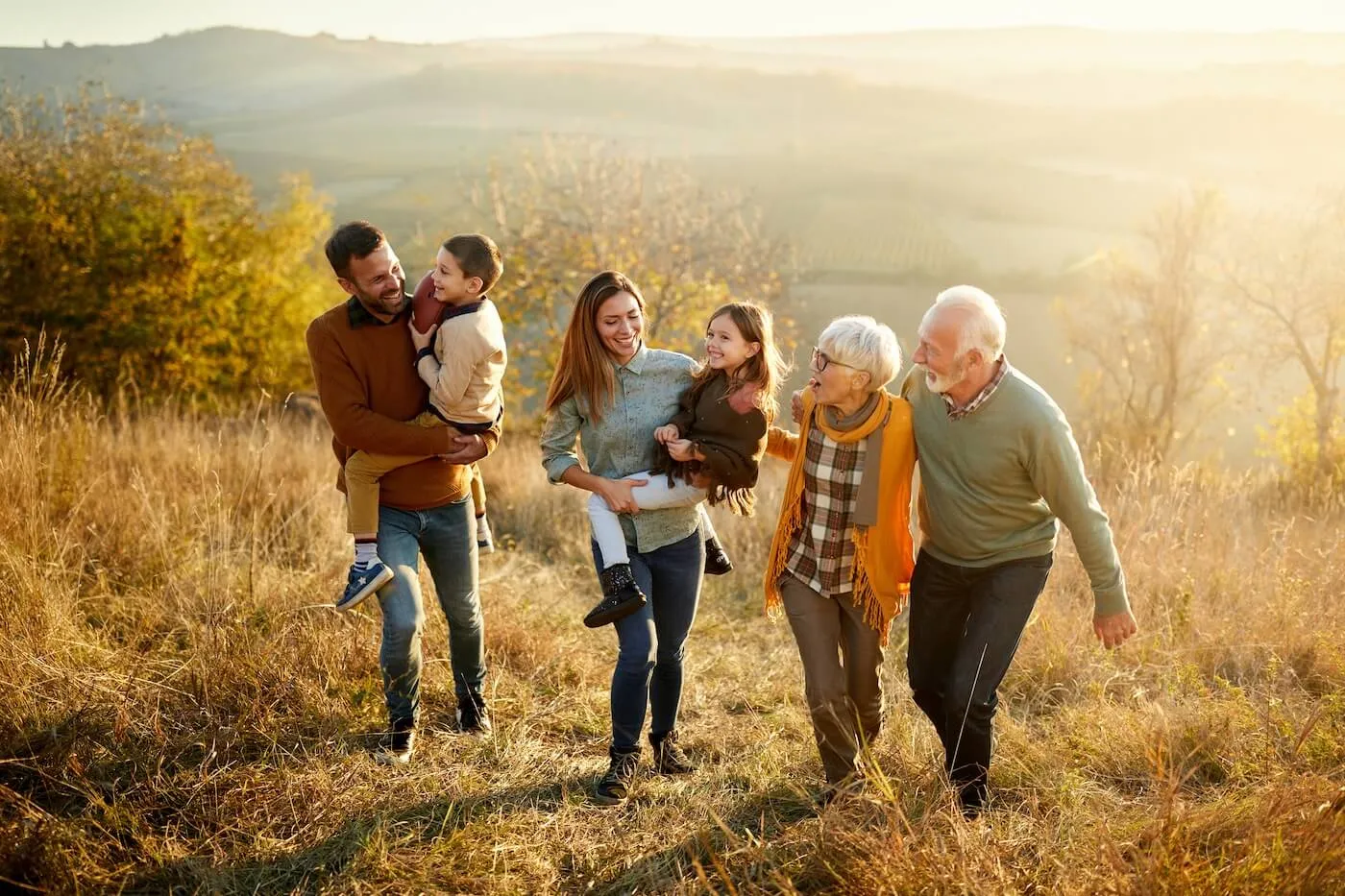 Multi-generational family walking outdoors on a sunny afternoon; parents carrying young children while grandparents stroll alongside, surrounded by nature