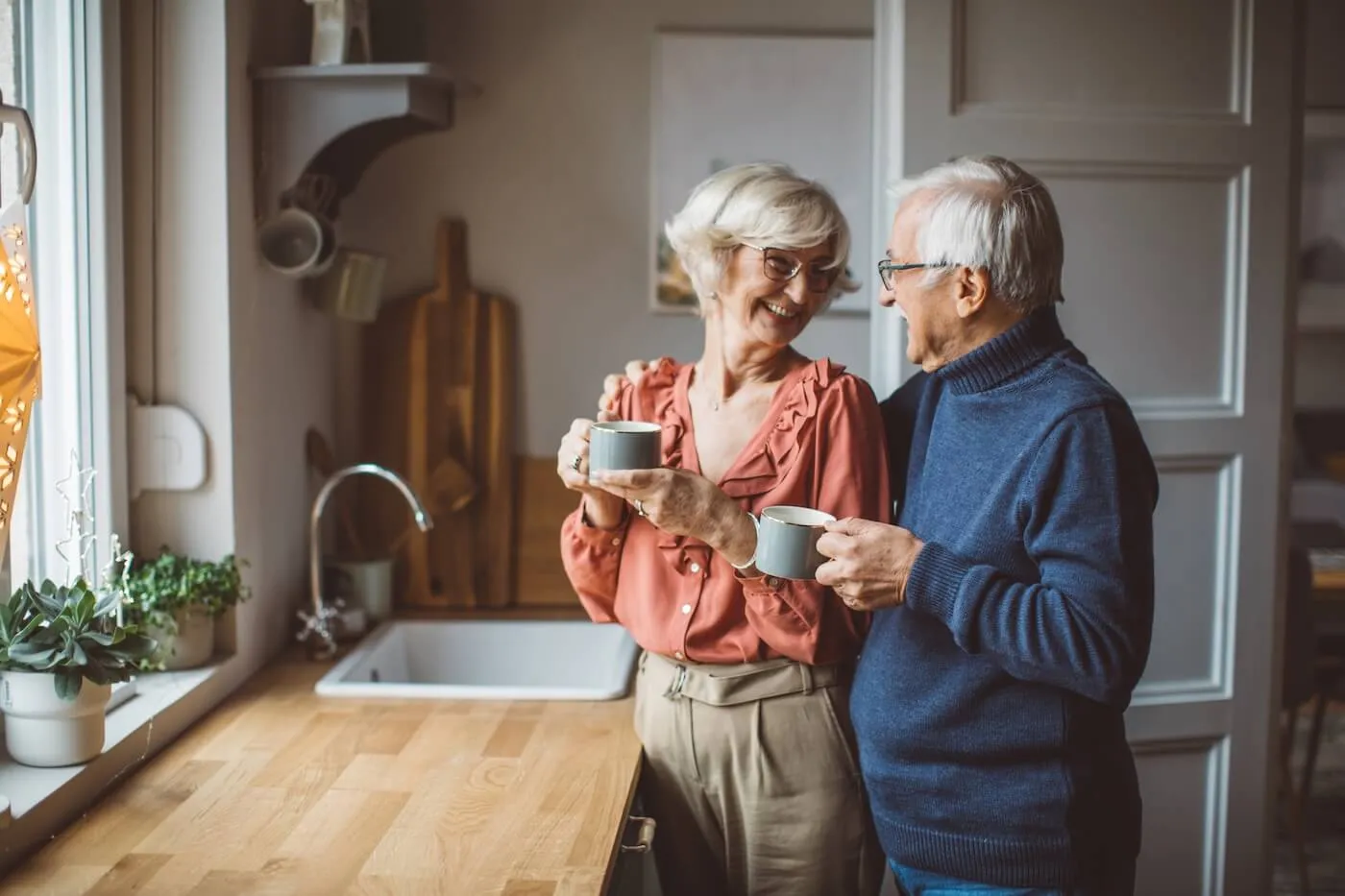 Happy older couple standing in the kitchen, hugging and holding mugs