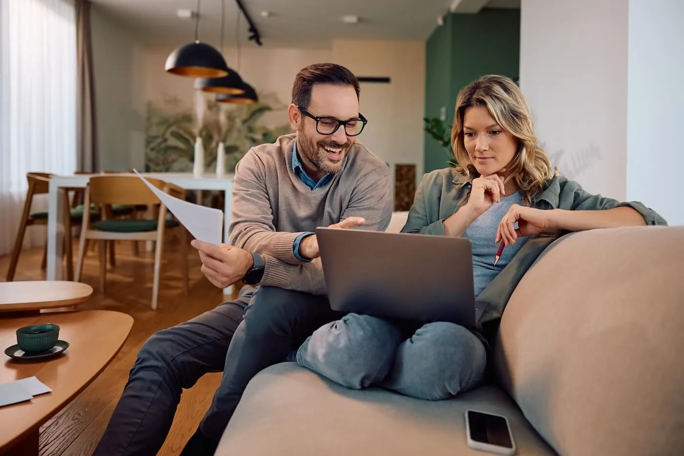 Smiling couple sitting on a sofa, reviewing savings account rates together; the man holds printed documents while pointing to something on the laptop screen for the woman.