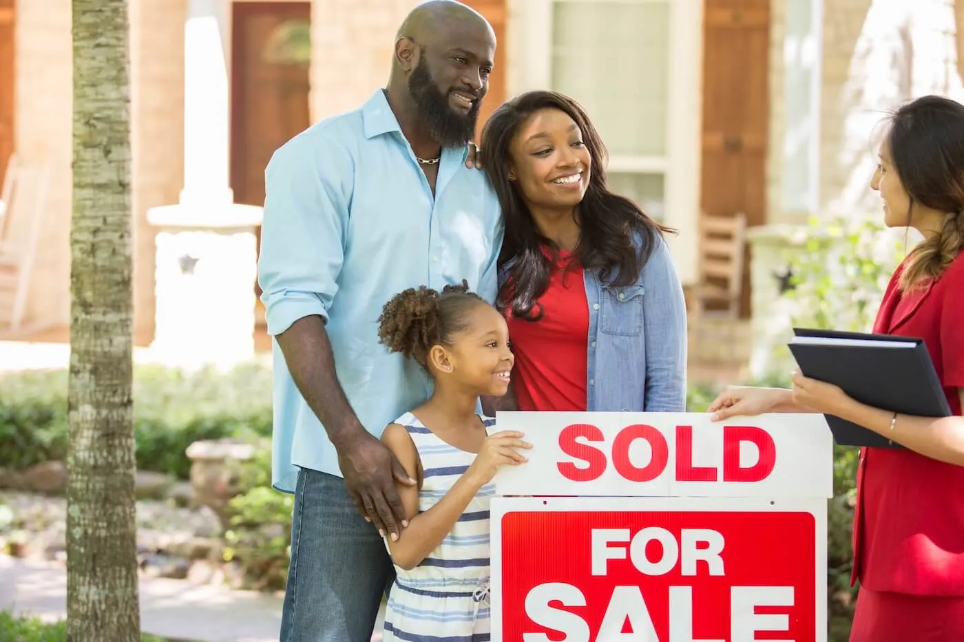 Smiling family of three standing with a female real estate agent in the front yard of their new home, as they place a SOLD sign on the lawn.