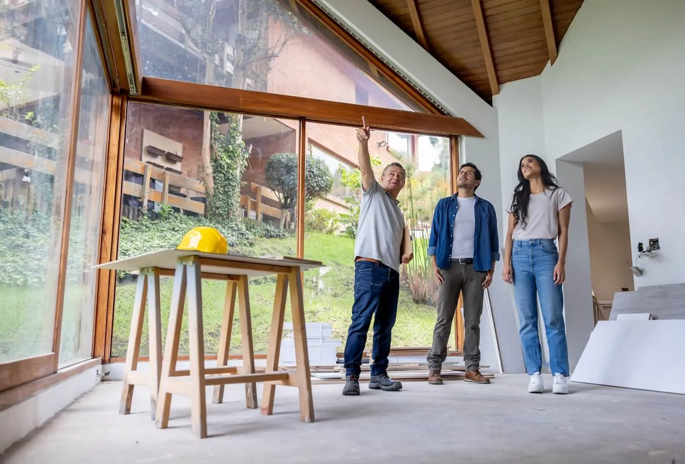 A young couple stands with a construction engineer in the middle of a partially renovated living room. They are engaged in a discussion, gesturing toward the ceiling as they assess the condition of the roof.