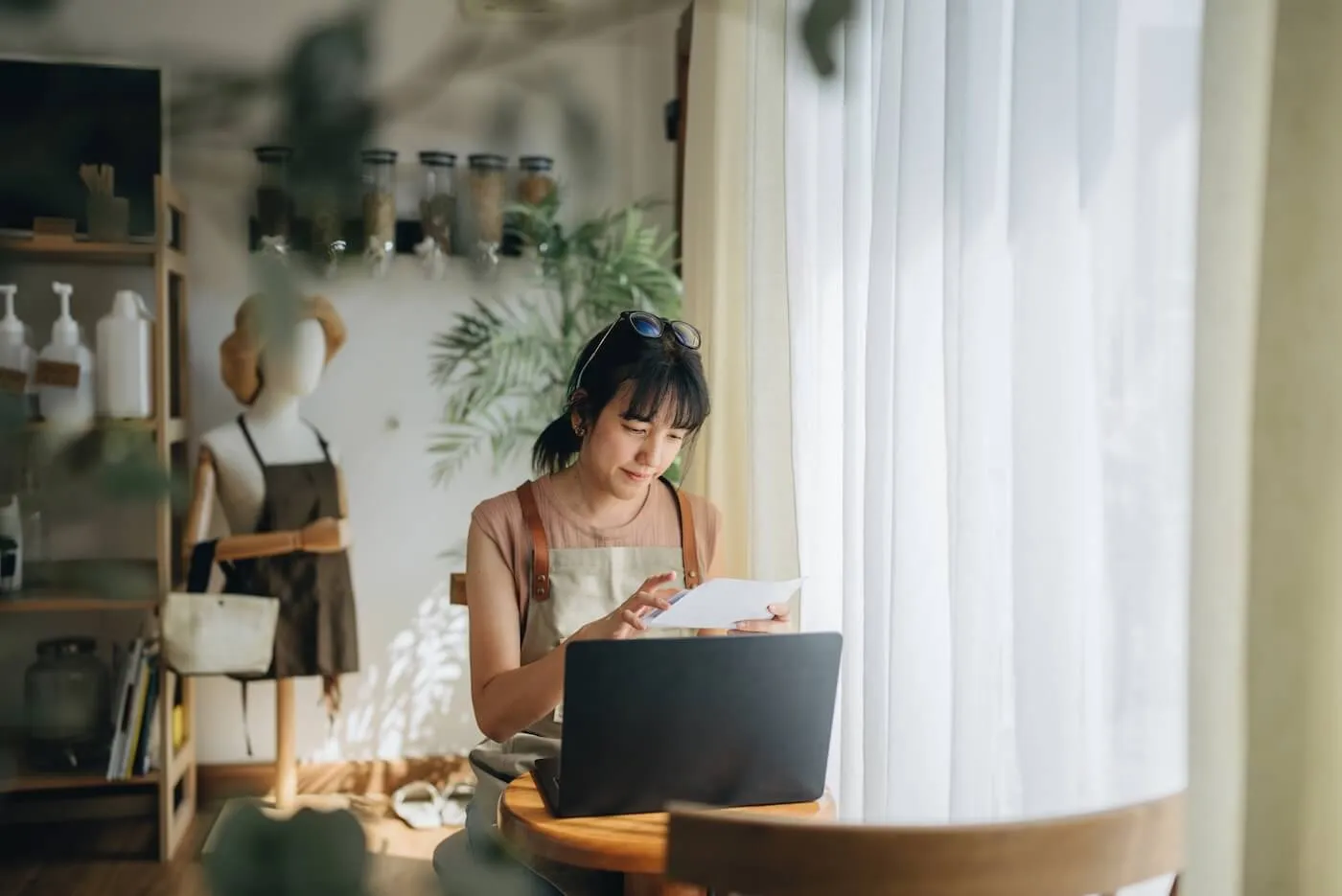A small business owner reviewing financial documents and using a laptop in her sunlit store, surrounded by shelves of products and plants