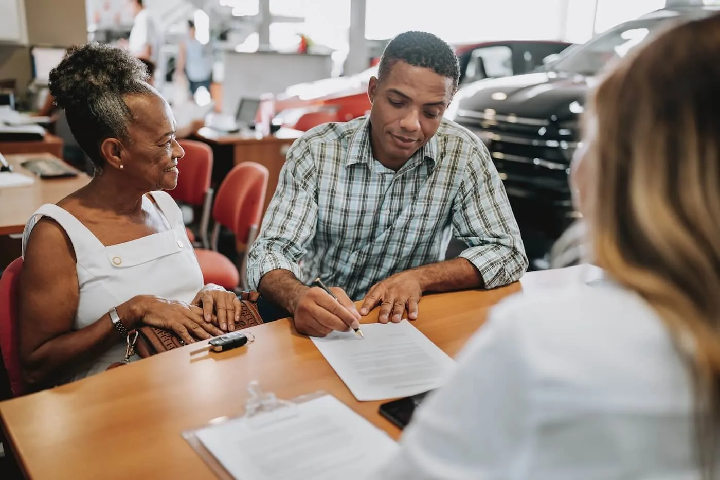 Young man signing the papers in a car dealership with a senior woman and a sales agent sitting next to him.