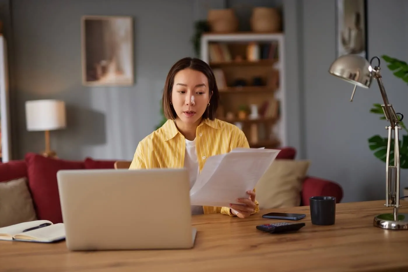 Focused young woman sits at a desk reviewing a document while using a laptop, with a phone, notebook, and calculator on the table in a home setting.