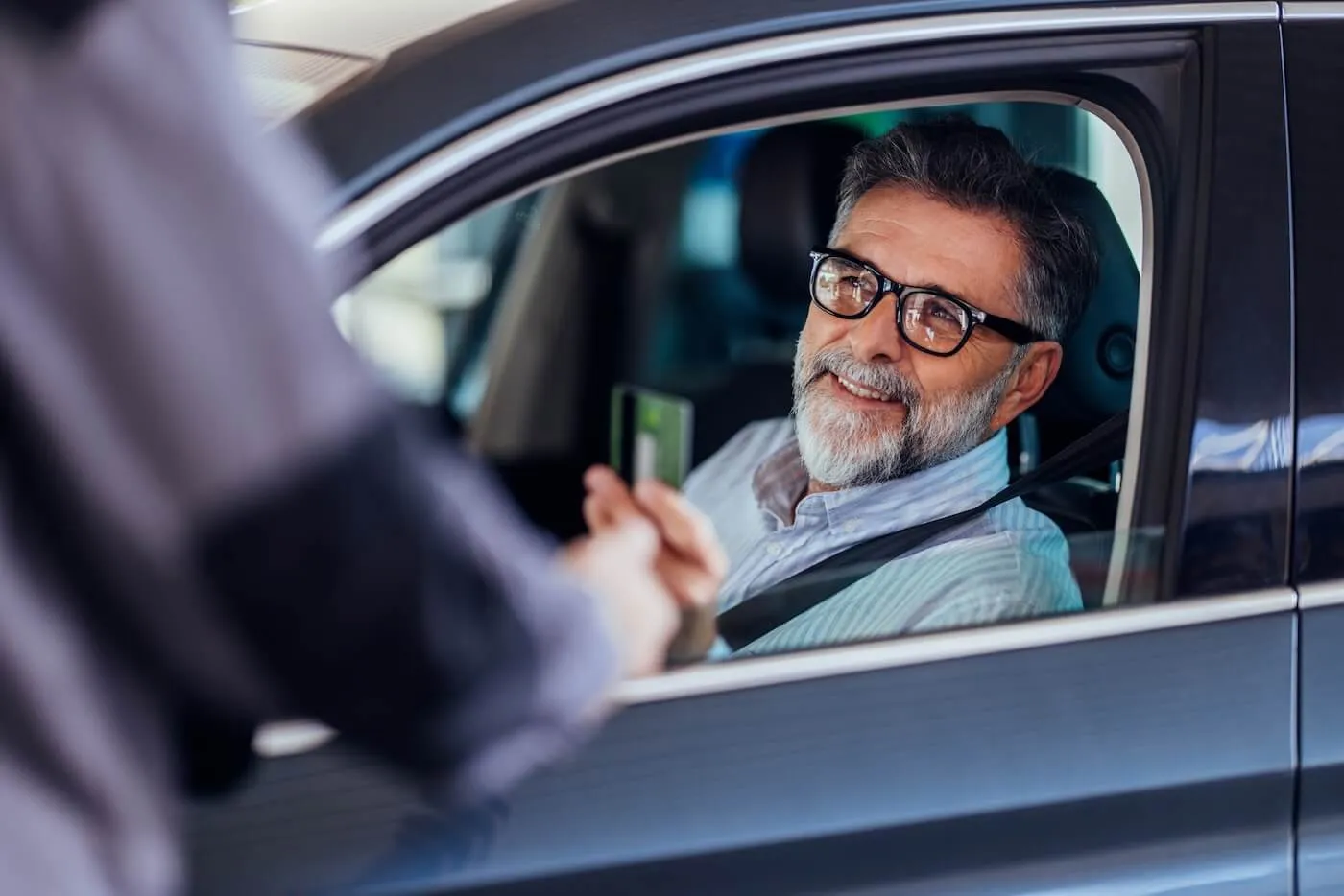 Smiling mature man wearing glasses, sitting in the driver’s seat, handing a credit card to someone through the car window