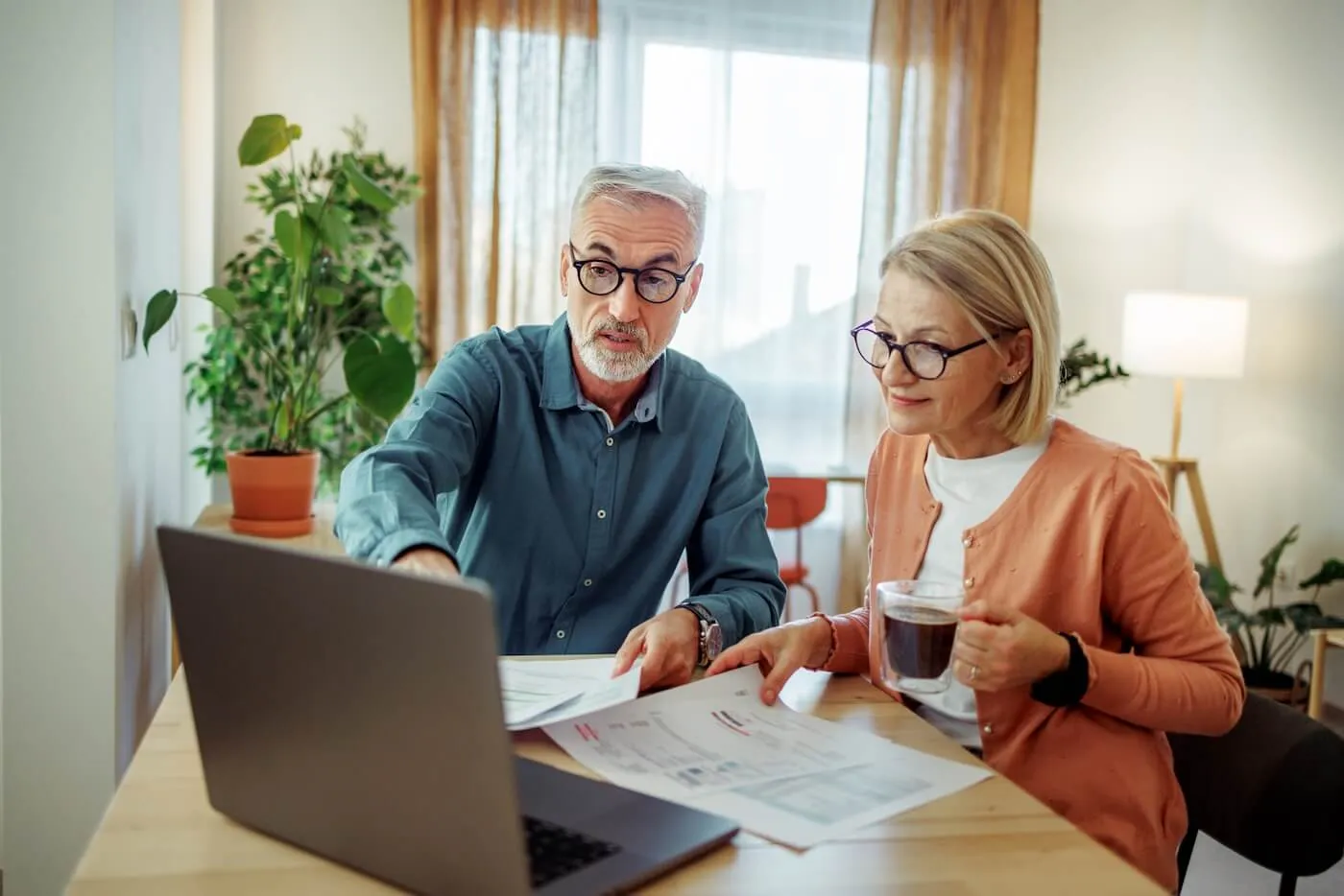 Mature couple reviewing their finances at home, surrounded by printed documents; the man points at the laptop screen while the woman holds a coffee mug.