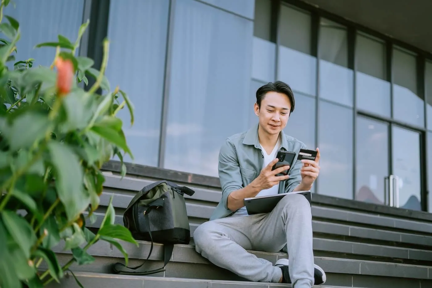 Smiling young man sitting on outdoor stairs with a tablet on his lap, using his mobile phone and holding a credit card, with a backpack placed beside him