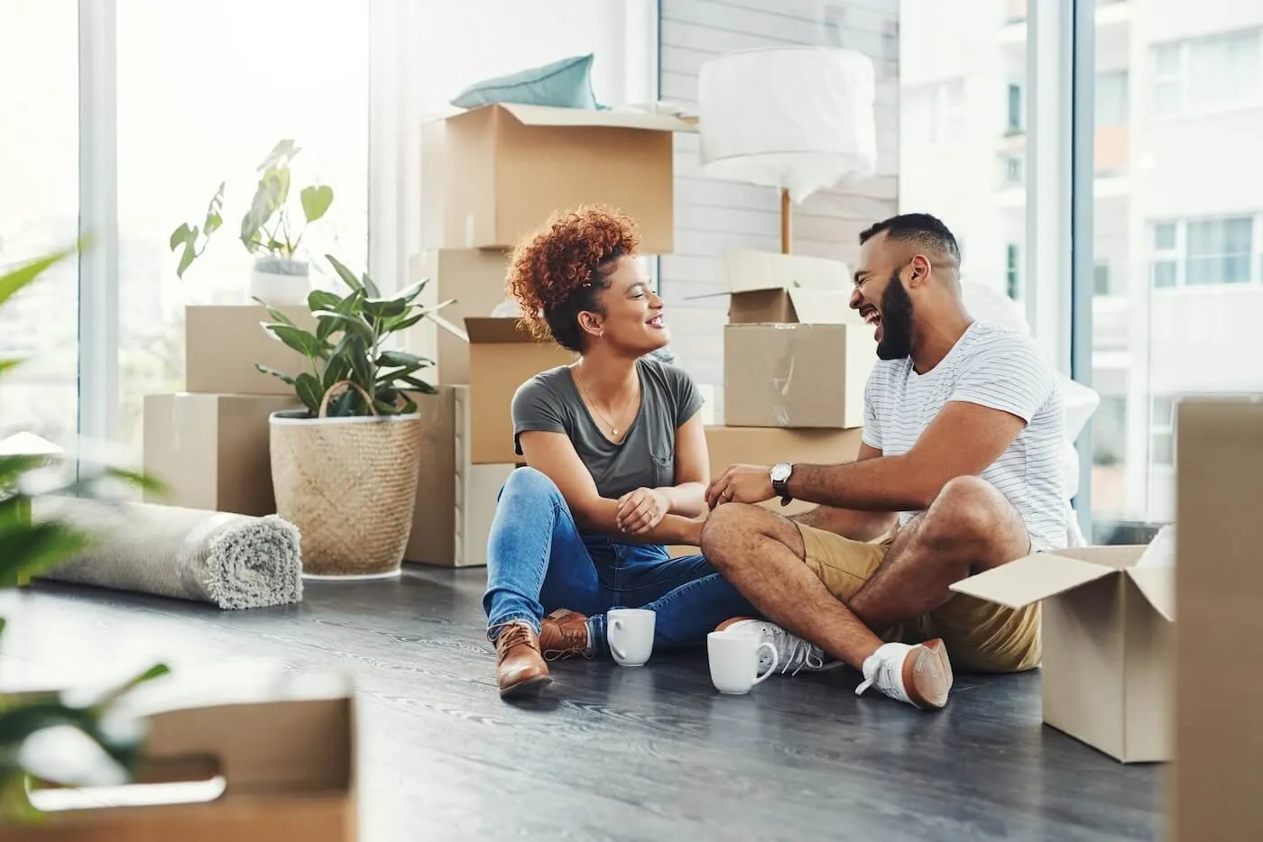 Smiling young couple sitting on the floor of their new apartment, surrounded by moving boxes, taking a break from unpacking and enjoying a hot drink