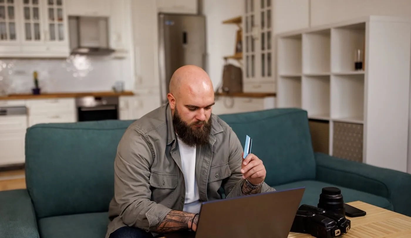 Male freelancer sitting on the sofa in the living room and using his credit card online, with a photo camera beside him
