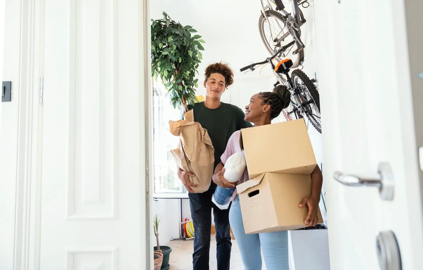 Young couple moving into a new apartment, carrying moving boxes and a plant, with a bicycle hanging on the wall behind them