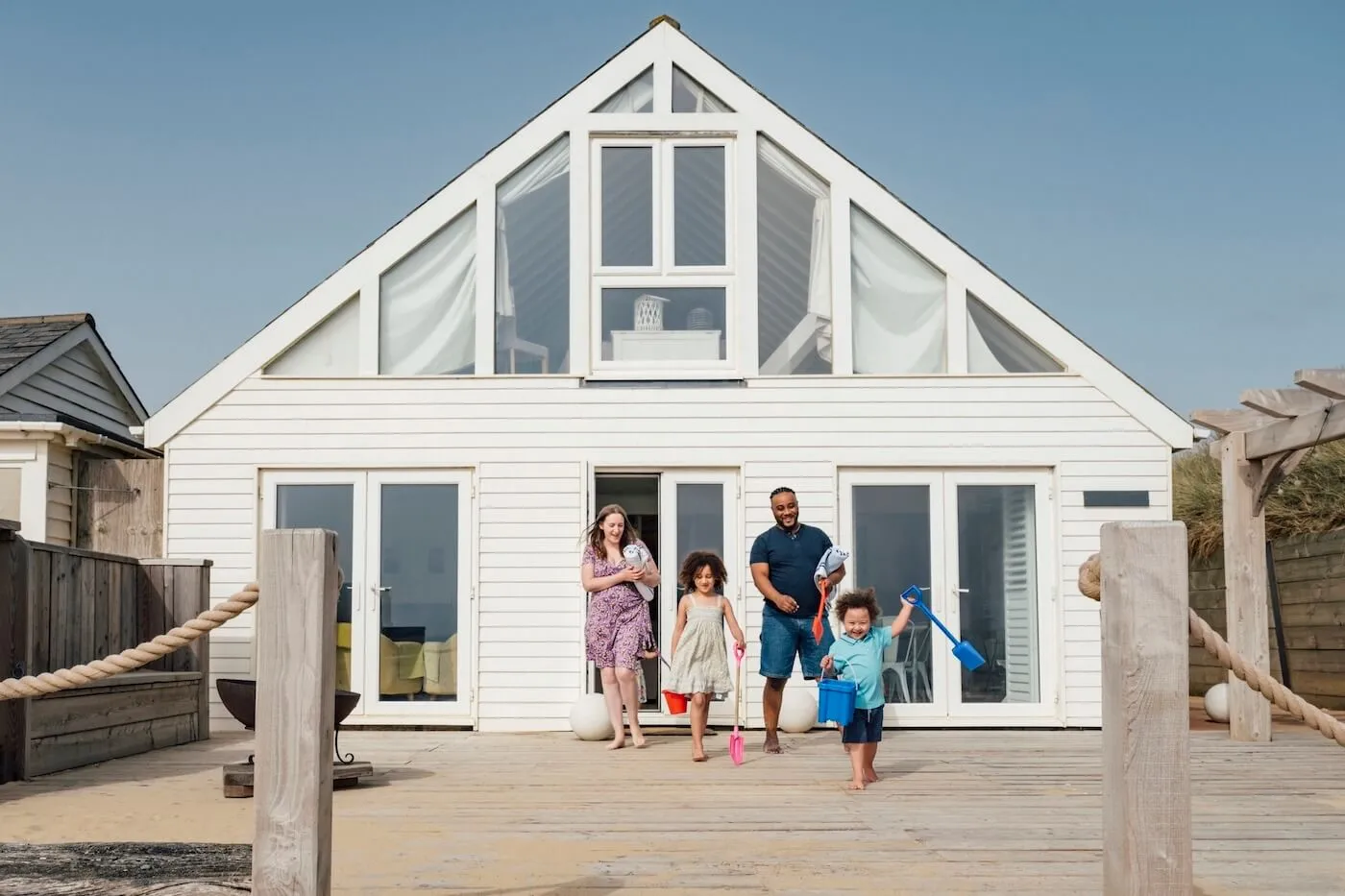 Happy family of four with two young kids leaving a white beach cottage on a sunny day