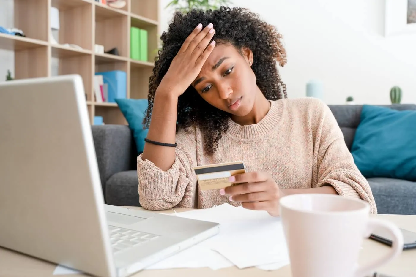 Concerned young woman looking at a credit card while using a laptop, with printed documents on the desk