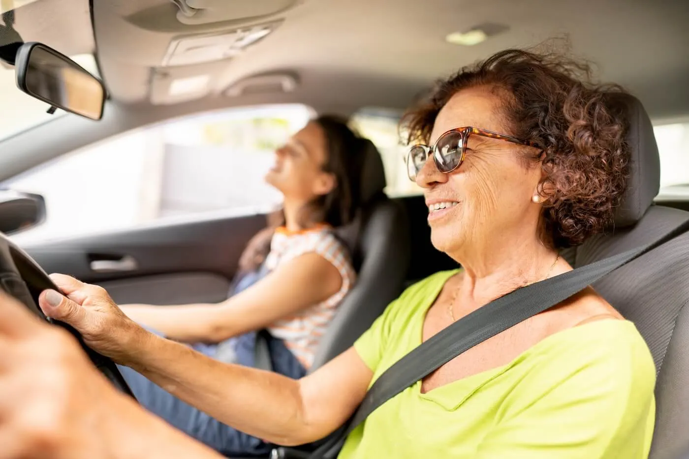 Smiling senior woman in sunglasses driving a car with a teenage girl on a passenger seat