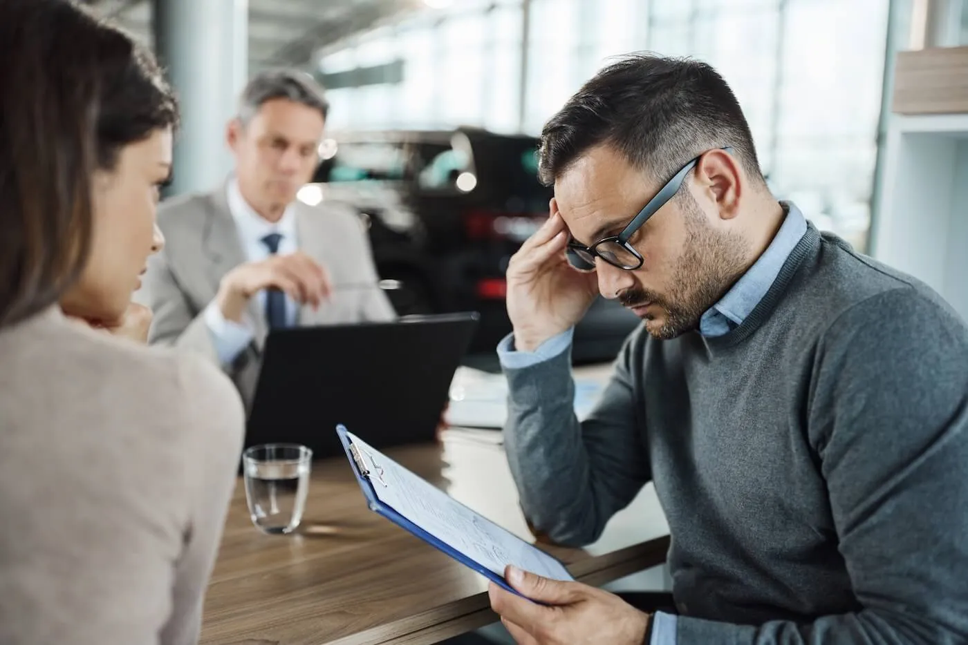 Concerned couple seated at a car dealership desk; the man is carefully reading a document while a male sales agent sits across from them, observing