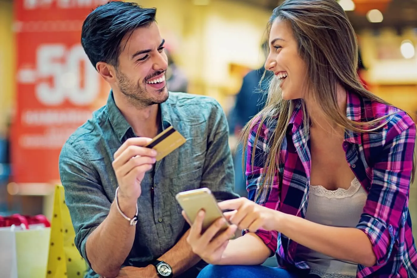 Happy young couple in a department store holding a credit card and a smartphone, with a sale poster in the background