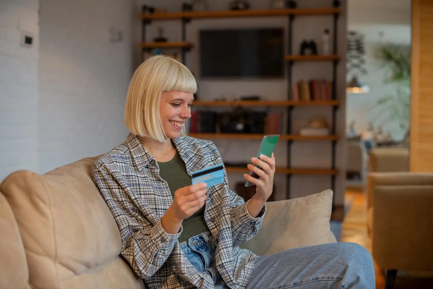 Smiling young woman sitting on a sofa and using her credit card in the mobile app