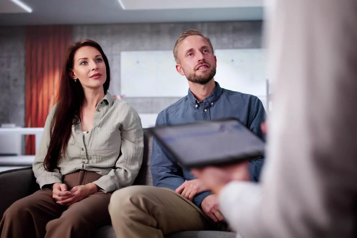 Two clients seated on a sofa during a consultation with a professional advisor in a modern office, while the advisor reviews information on a tablet.