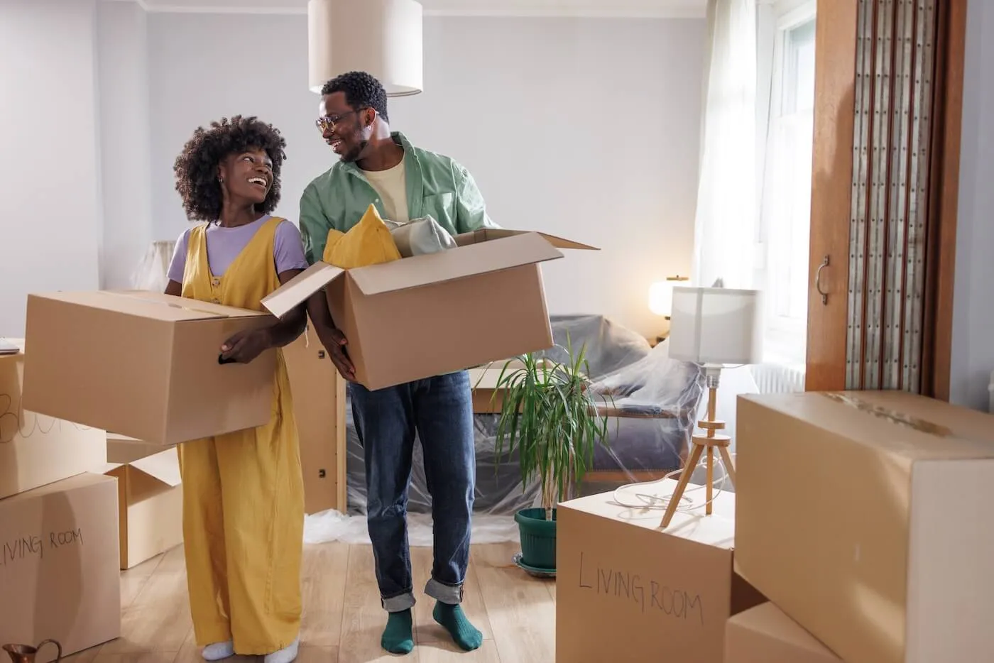 Smiling couple carrying moving boxes into their new home, surrounded by unpacked items