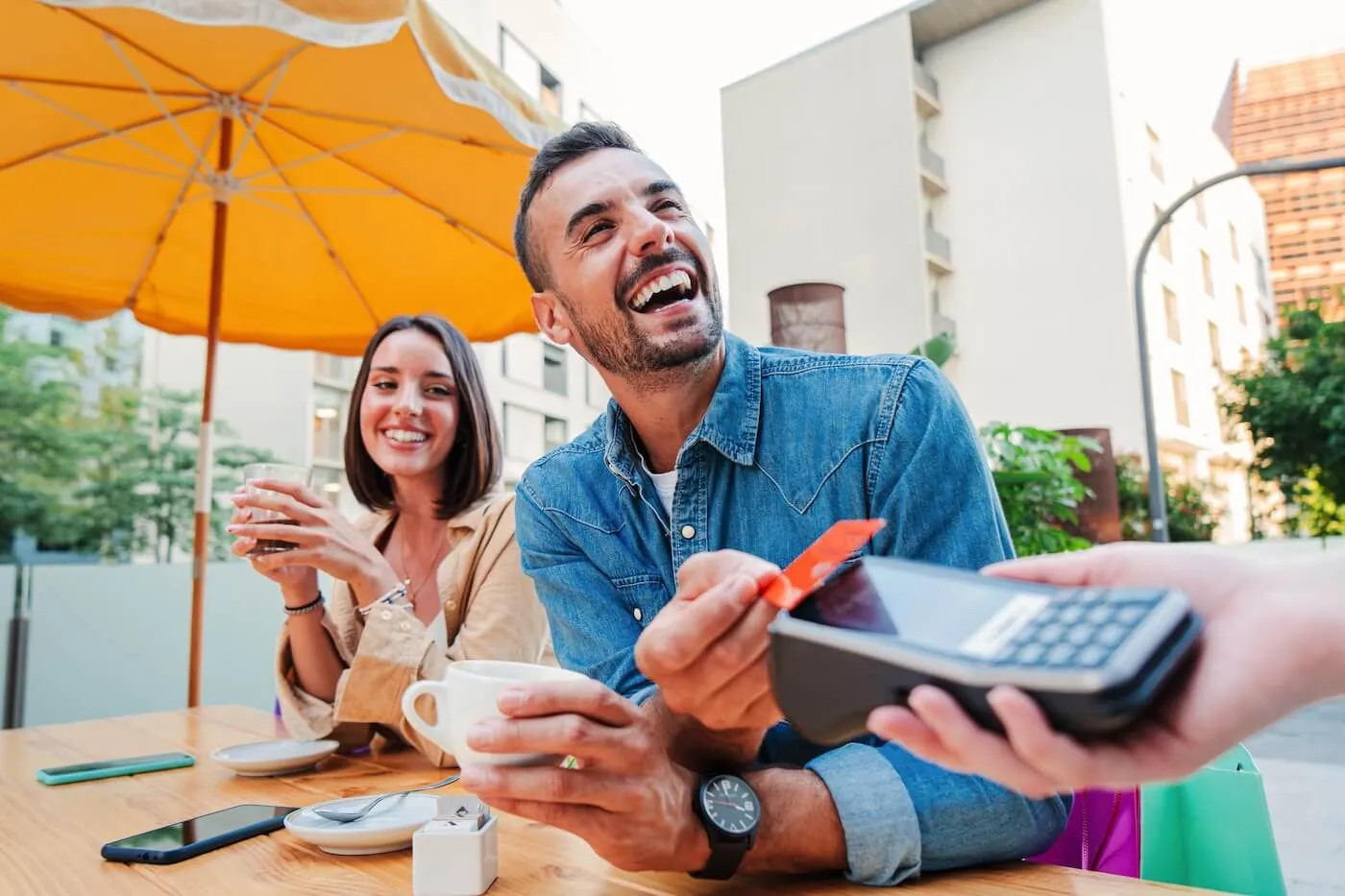 Smiling man making a contactless payment with a credit card at an outdoor café table while holding a coffee cup, with a payment terminal extended by a server and another café guest seated nearby.