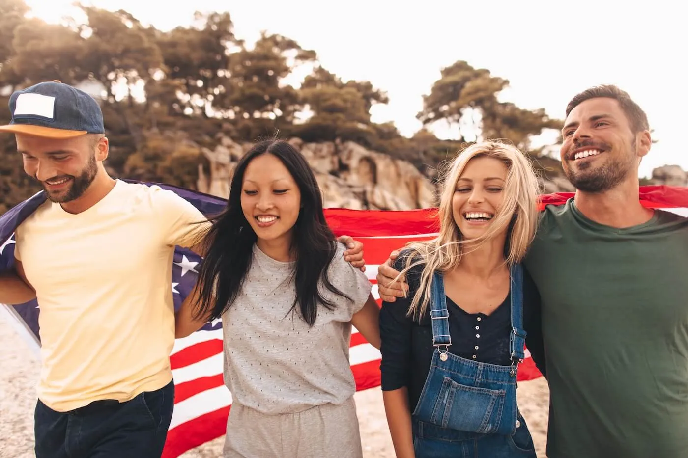 A group of multinational friends standing side by side with their arms around each other, holding an American flag draped across their shoulders. They are outdoors in a natural setting with trees and rocky terrain in the background.