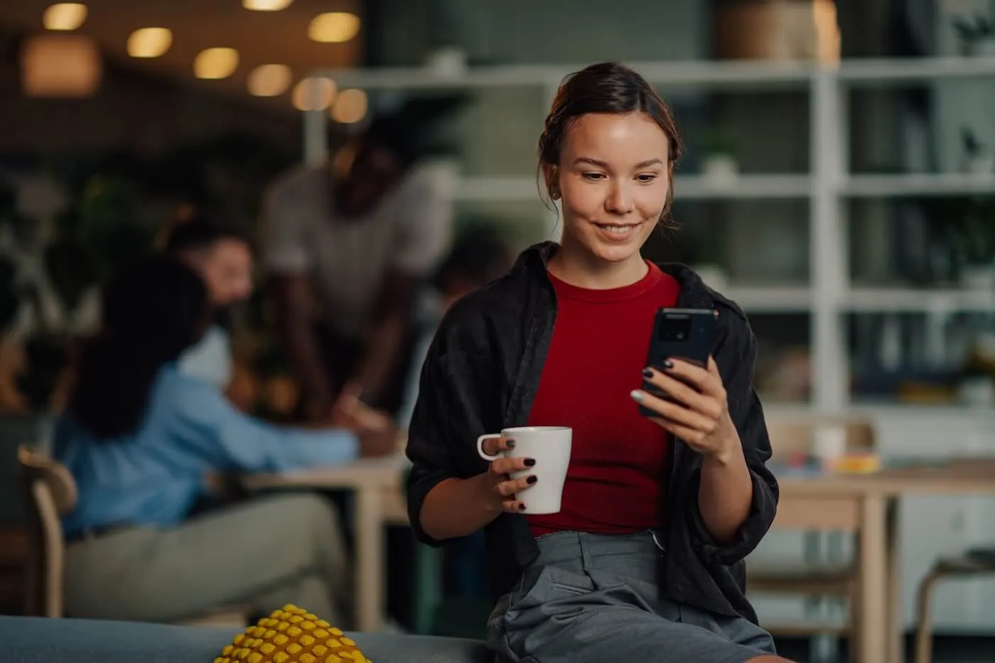 Smiling young woman on a sofa armrest using her phone and holding a mug in a busy café
