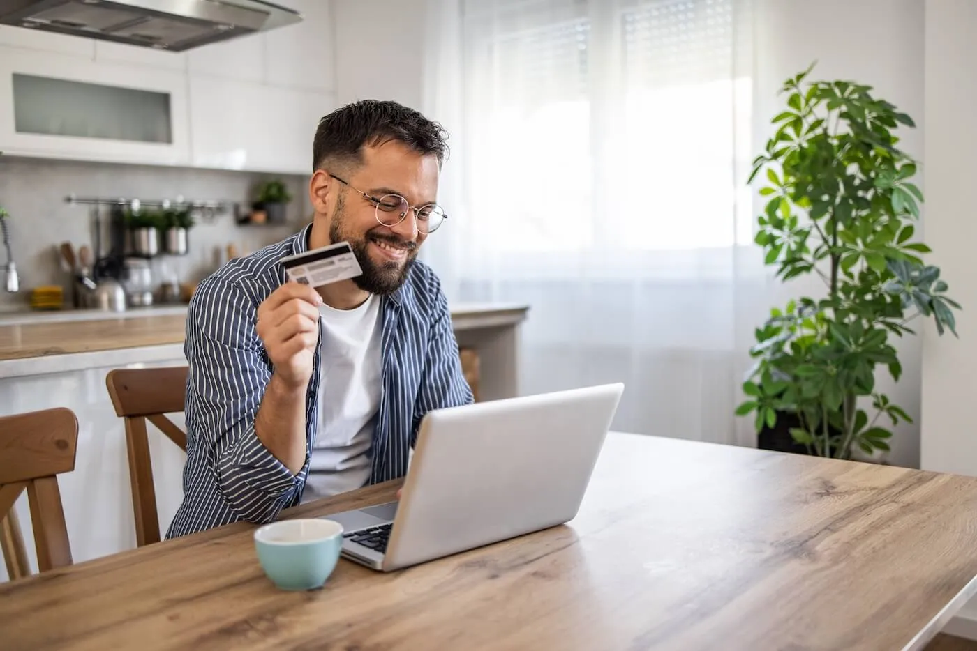 Smiling man wearing glasses is using his credit card online in the kitchen
