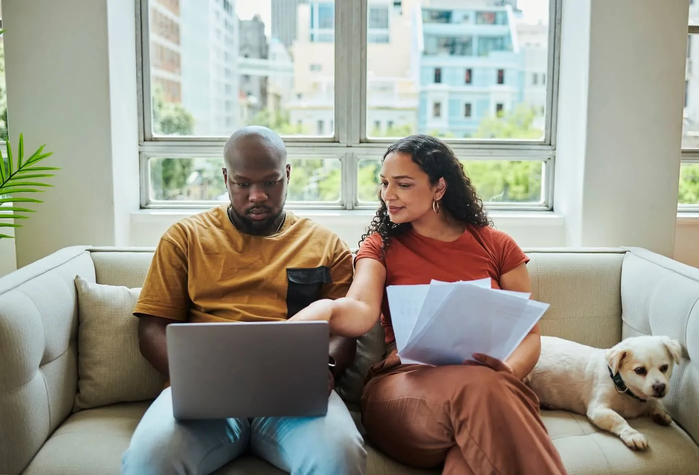 Young couple sitting on the sofa with a laptop and a small dog; the woman is holding some printouts and pointing at the laptop screen