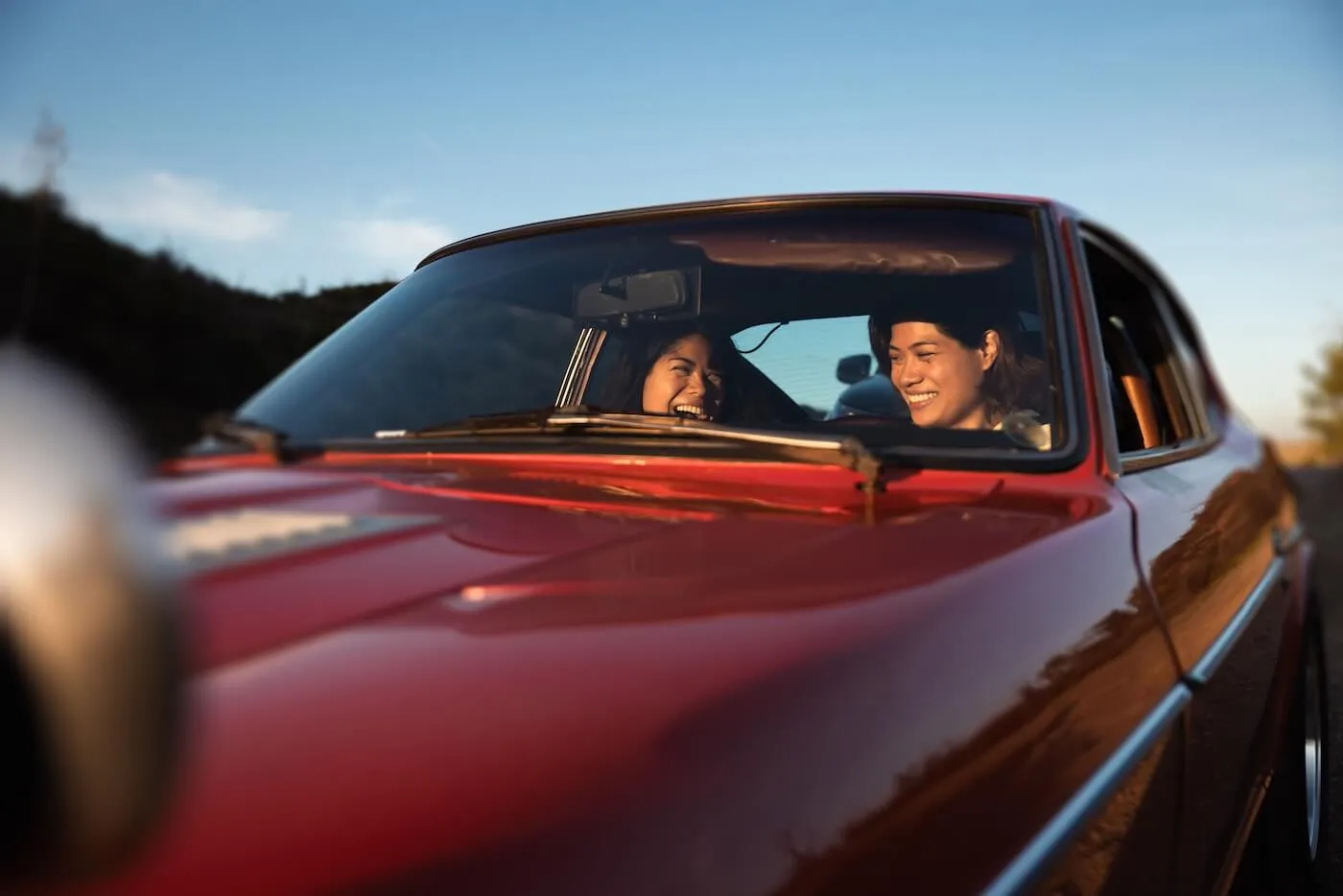 Two happy female friends sitting inside a red car, viewed from the front through the windshield, both look toward each other while traveling on a road, with warm sunlight reflecting off the car’s hood and a natural landscape visible in the background
